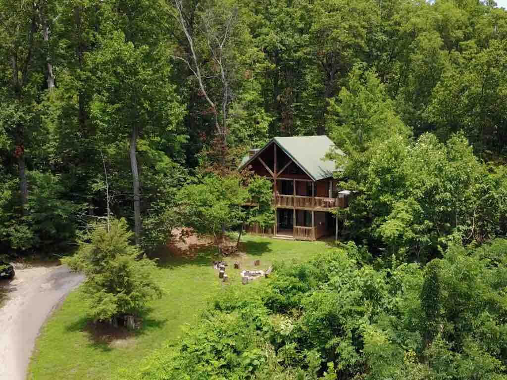 Aerial view of Thistle Britches log cabin with green roof on a private acre surrounded by Smoky Mountain forest, fire pit and gravel driveway visible